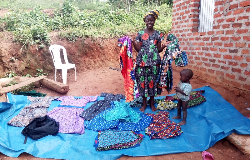 Lucy stands on a tarpaulin sheet holding dresses she has made in each hand, and displaying other dresses on the sheet