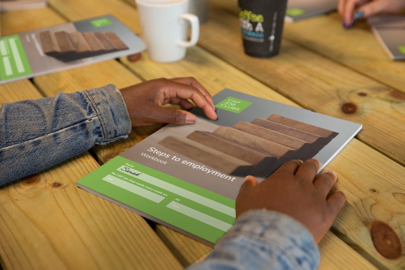 Shot of a man's hands on a table, holding a CAP Job Club document stating "Steps to employment workbook"