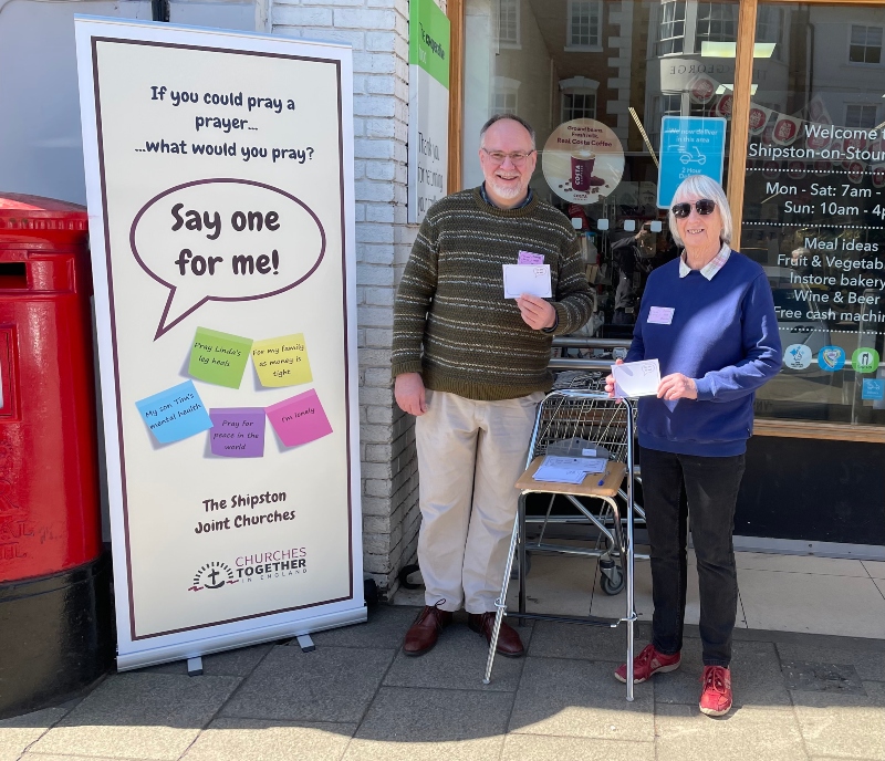 A man and woman stand outside a coffee shop holding cards; they are next to a banner which says "Say one for me!"