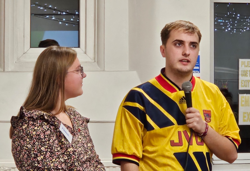 Youth Alpha testimony - a young man in a yellow t-shirt speaks while holding a microphone. He is stood next to Hannah, a Youth Alpha host