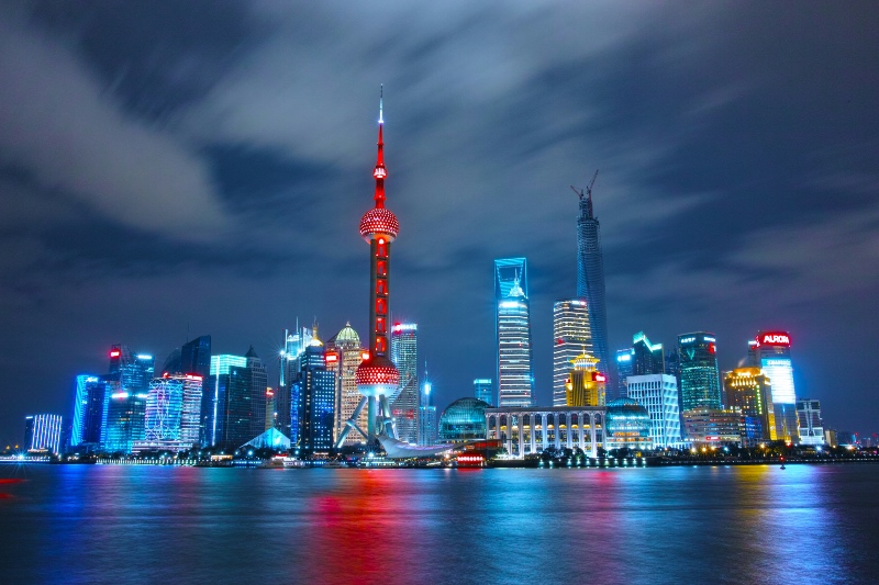 Shanghai skyline at night, featuring brightly lit skyscrapers reflected on the river. The iconic Oriental Pearl Tower glows in red amid a cloudy sky