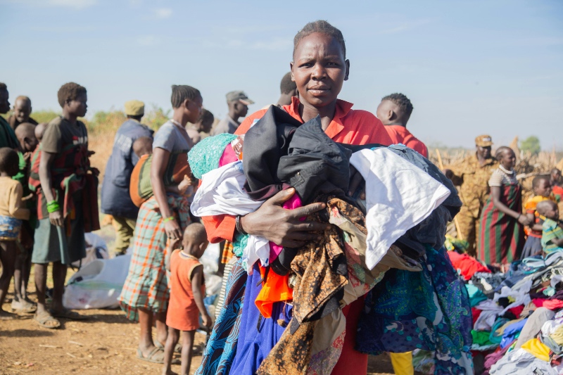 A woman in South Sudan stands outdoors, holding a bundle of clothes, surrounded by others in colorful attire, some seated on the ground.