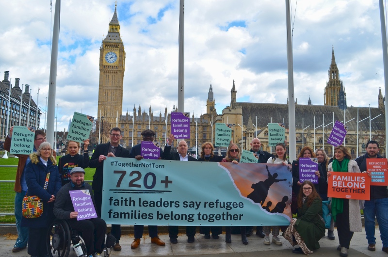 A diverse group holds a banner reading "720+ faith leaders say refugee families belong together" outside UK Parliament. Signs state "Refugee families belong together". Big Ben is visible, under a cloudy sky.