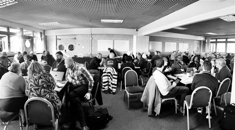 A black and white image showing a busy conference environment with people engaged in discussions around tables. The room is filled with natural light.