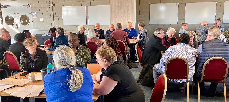 A diverse group of people sit at round tables in a well-lit room, engaged in focused discussion