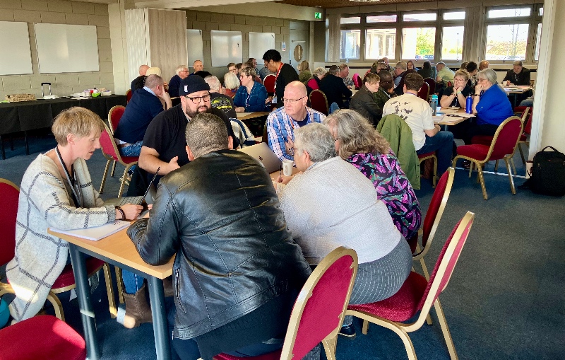A group of people seated in clusters around tables in a bright meeting room. They are engaged in discussions, creating a collaborative and focused atmosphere.