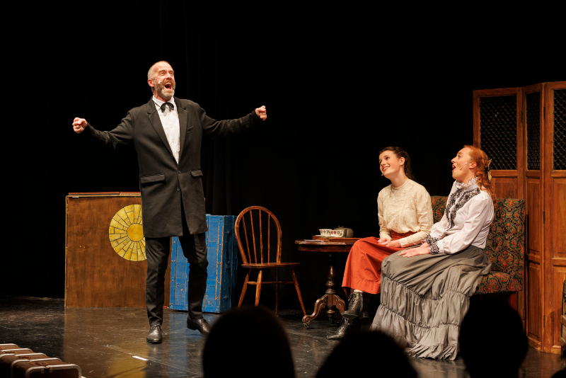 A man in a black coat energetically gestures on stage, while two seated women in period costumes smile and engage. The setting is a cozy, vintage room.
