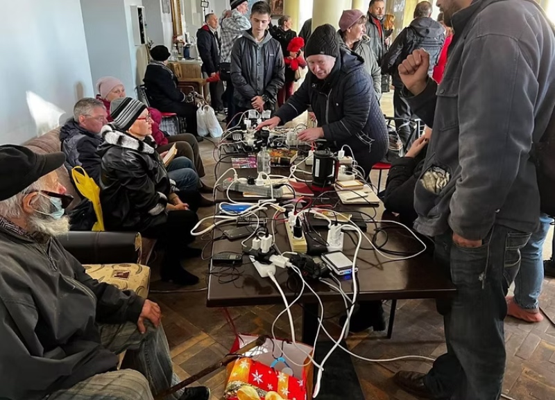 A group of people gathered around long tables filled with power strips, cables, and charging electronic devices inside a community space.