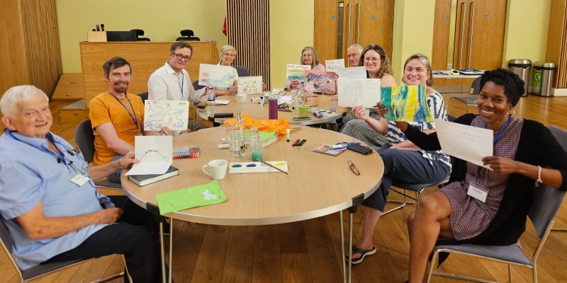 A group of people seated around wooden tables indoors, smiling and holding up colourful drawings and artwork created during the Small Church Connexion retreat
