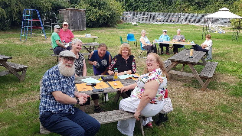 A group of people sitting around picnic tables during an outdoor gathering on a grassy field.