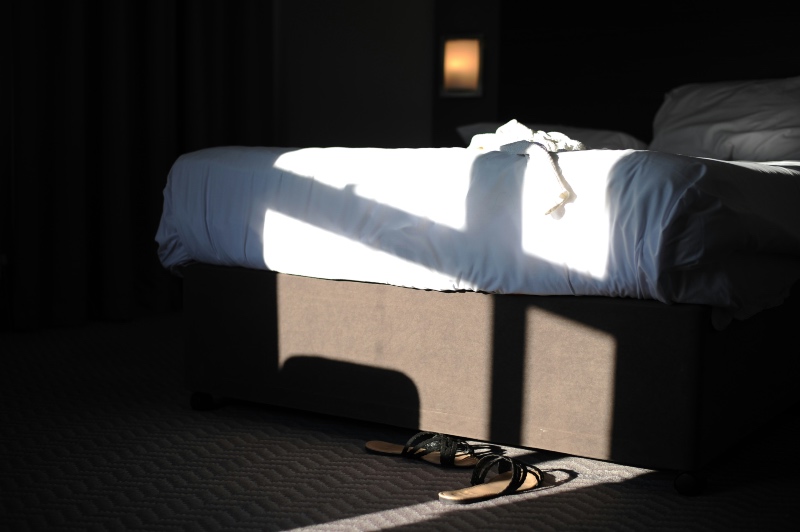 A bed in a dimly lit room with bright sunlight streaming across the white duvet and a pair of sandals on the floor beside it.