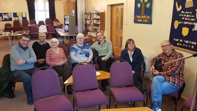 A group of seven people sitting in a semi-circle of chairs inside a church hall 