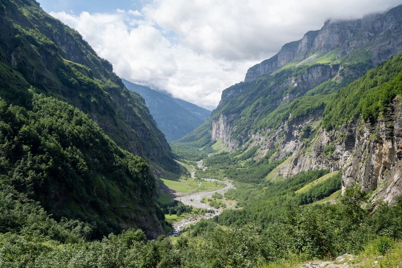 A wide view looking down a dramatic mountain valley framed by steep, forest-covered slopes and rocky cliffs under a partly cloudy sky