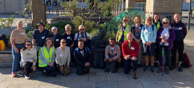 A large group of walkers posing together outdoors in a sunny public square, with some members wearing high-visibility vests or holding walking poles